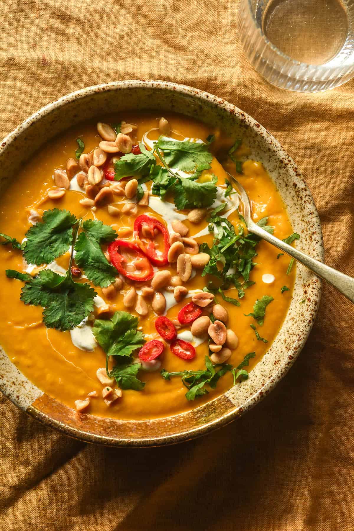 An aerial image of a bowl of low FODMAP Thai pumpkin soup topped with chillies, coconut cream, coriander and peanuts. The bowl sits on a mustard coloured linen tablecloth and a glass of water sits to the top right of the image.