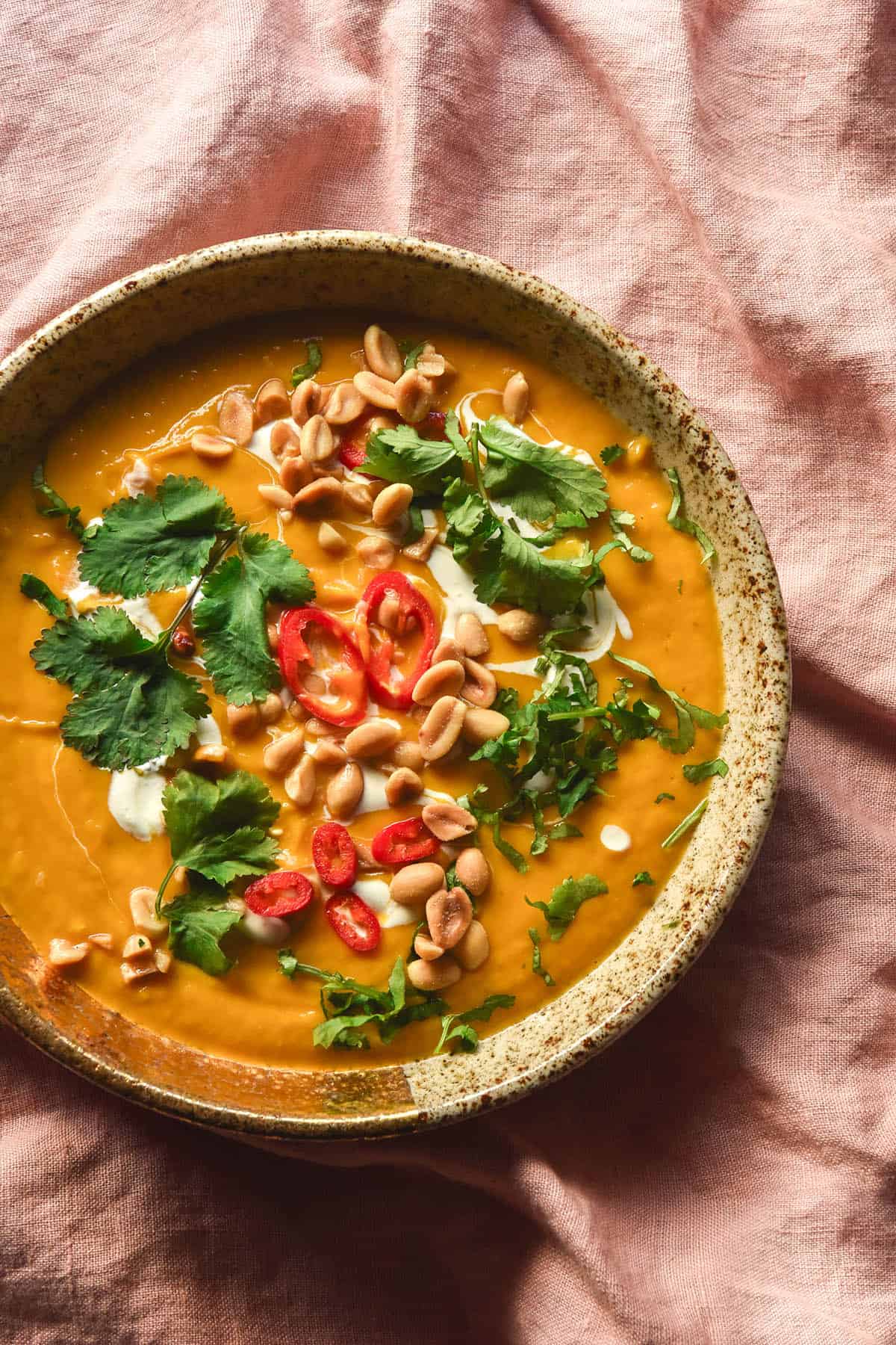 An aerial image of a bowl of low FODMAP Thai pumpkin soup topped with chillies, coconut cream, coriander and peanuts. The bowl sits on a casually strewn pale pink linen tablecloth.