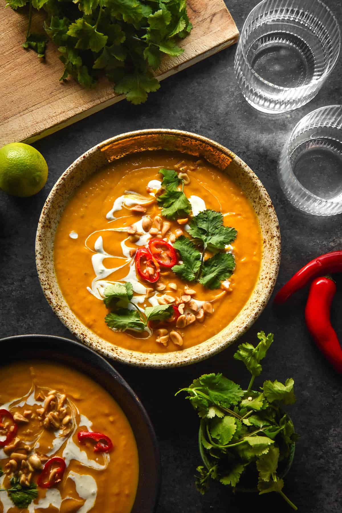 An aerial image of a bowl of low FODMAP Thai pumpkin soup topped with chillies, coconut cream, coriander and peanuts. The bowl is surrounded by ingredients for the soup on a dark blue backdrop.