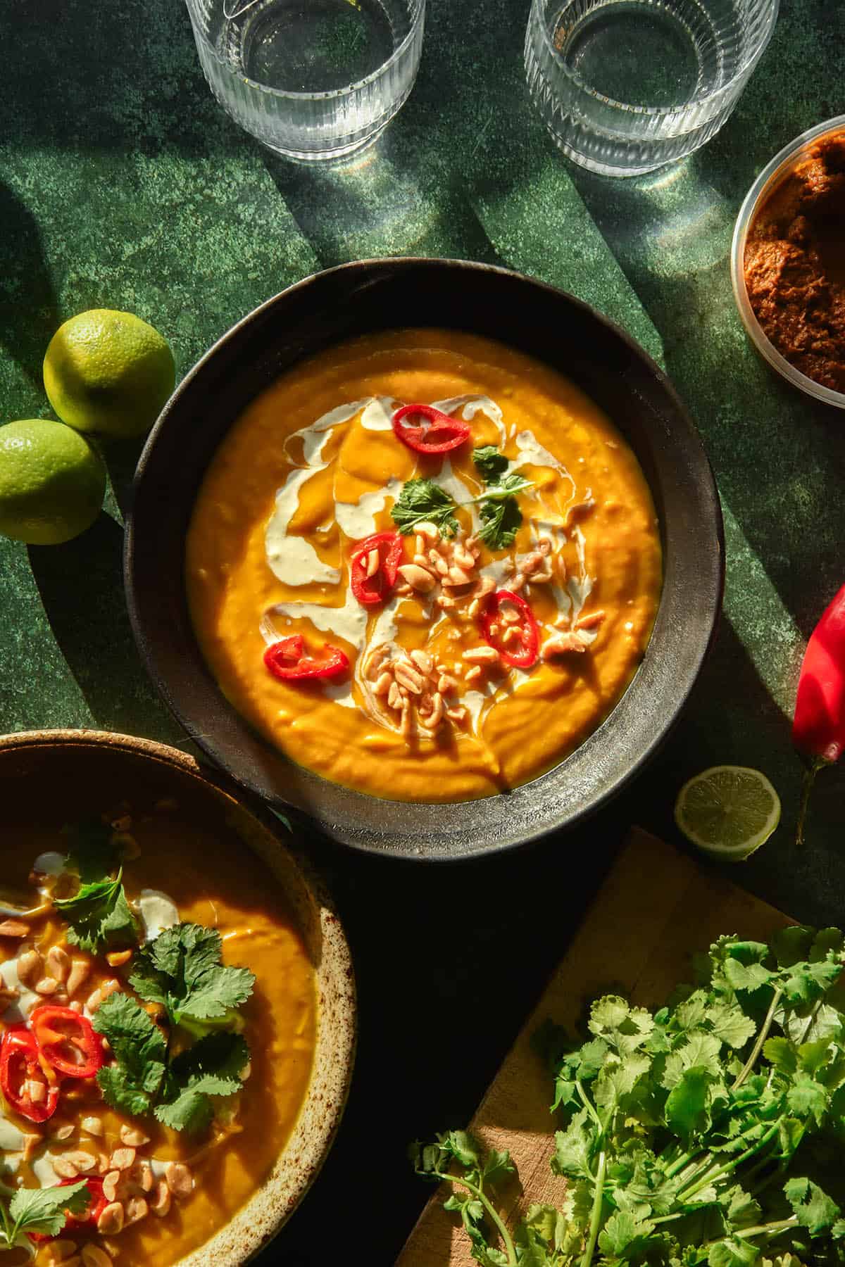 An aerial image of a bowl of low FODMAP Thai pumpkin soup topped with chillies, coconut cream, coriander and peanuts. The bowl sits on a dark green backdrop surrounded by ingredients for the soup and another bowl of soup in the bottom left corner.