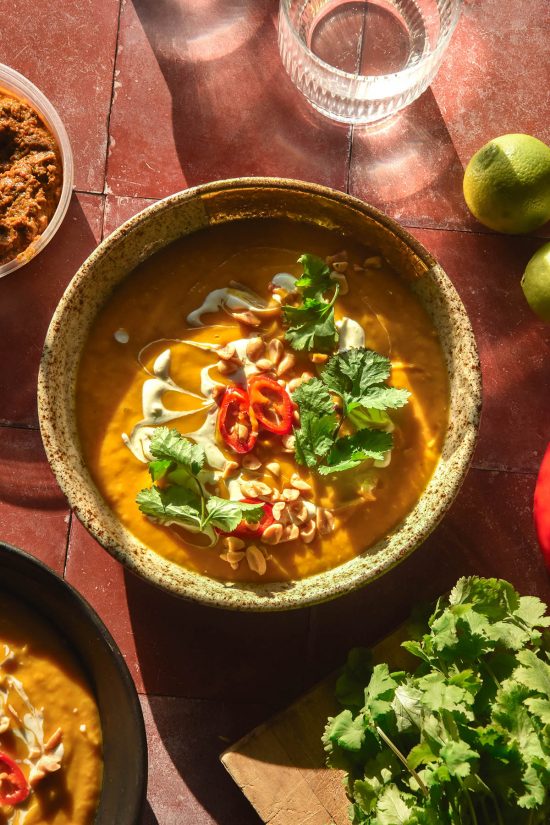 A brightly lit aerial image of a speckled ceramic bowl filled with low FODMAP Thai pumpkin soup. The soup is topped with coconut cream, chillies, coriander and peanuts and sits atop a terracotta tile backdrop. The bowl is surrounded by extra limes, chilli, coriander and sunlit glasses of water.