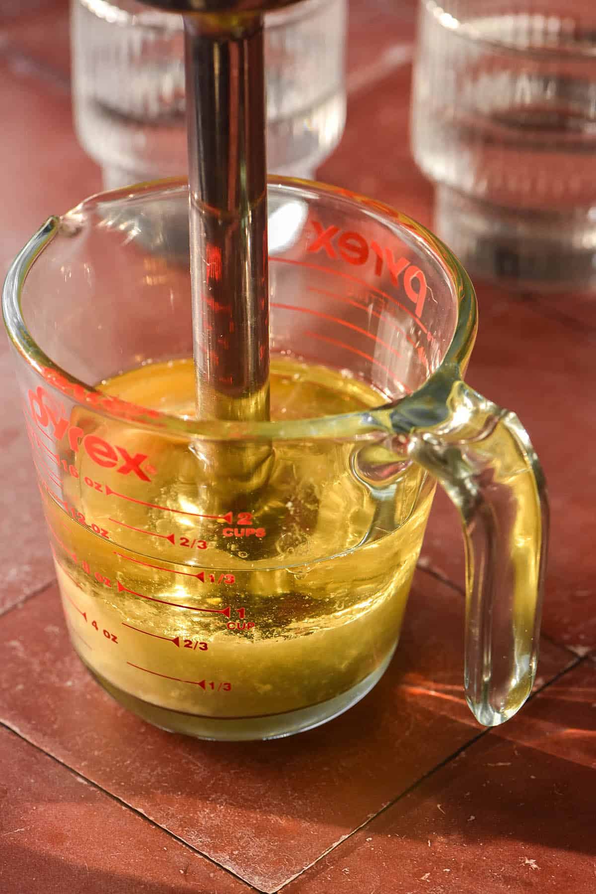 An image of a pouring jug filled with the ingredients for low FODMAP mayonnaise. The jug sits on a terracotta stone backdrop with an immersion blender sitting in the jug. Two glasses of water sit to the back right of the image.