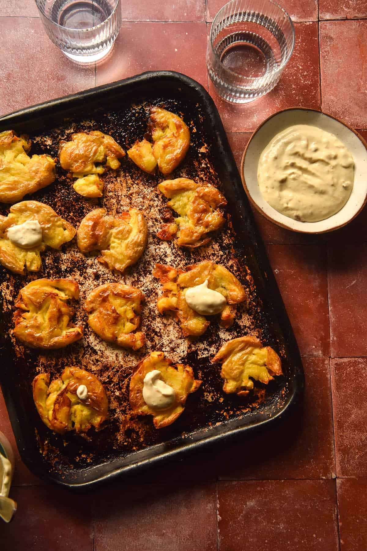 An aerial view of a tray of crispy roast smashed potatoes on a terracotta tile backdrop. A white bowl of low FODMAP aioli sits to the top right of the tray along with two sunlit water glasses.