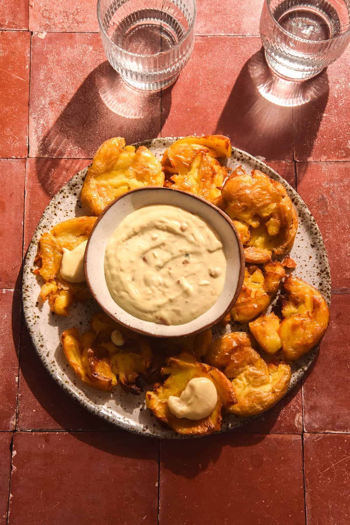 An aerial brightly lit image of a white ceramic plate topped with a bowl of low FODMAP aioli and surrounded by crispy smashed potatoes. Two sunlit glasses of water sit to the top of the image which is set atop a terracotta tile backdrop