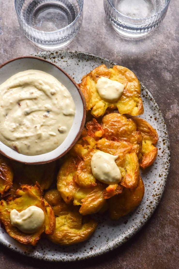 An aerial image of a plate of crispy smashed potatoes surrounding a bowl of Low FODMAP Garlic aioli. The plate sits atop a grey stone backdrop and two glasses of water sit the top of the image.