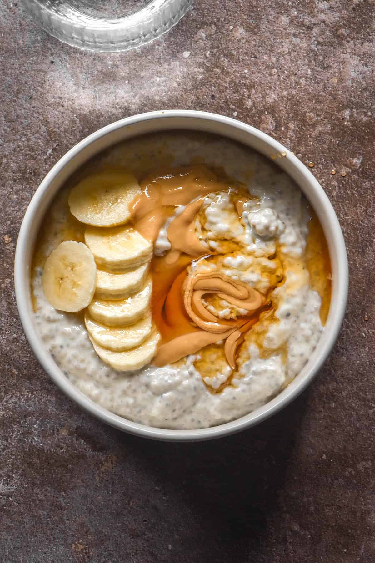 An aerial image of a white bowl of low FODMAP chia pudding topped with banana slices, peanut butter and maple syrup. The bowl sits atop a grey concrete backdrop and a glass of water sits to the top left of the image
