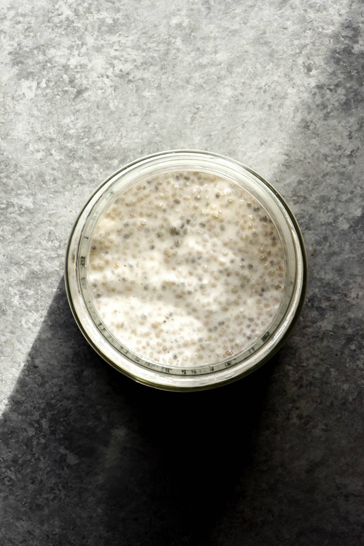 An aerial image of chia pudding in a jar atop a concrete background in contrasting sunlight.