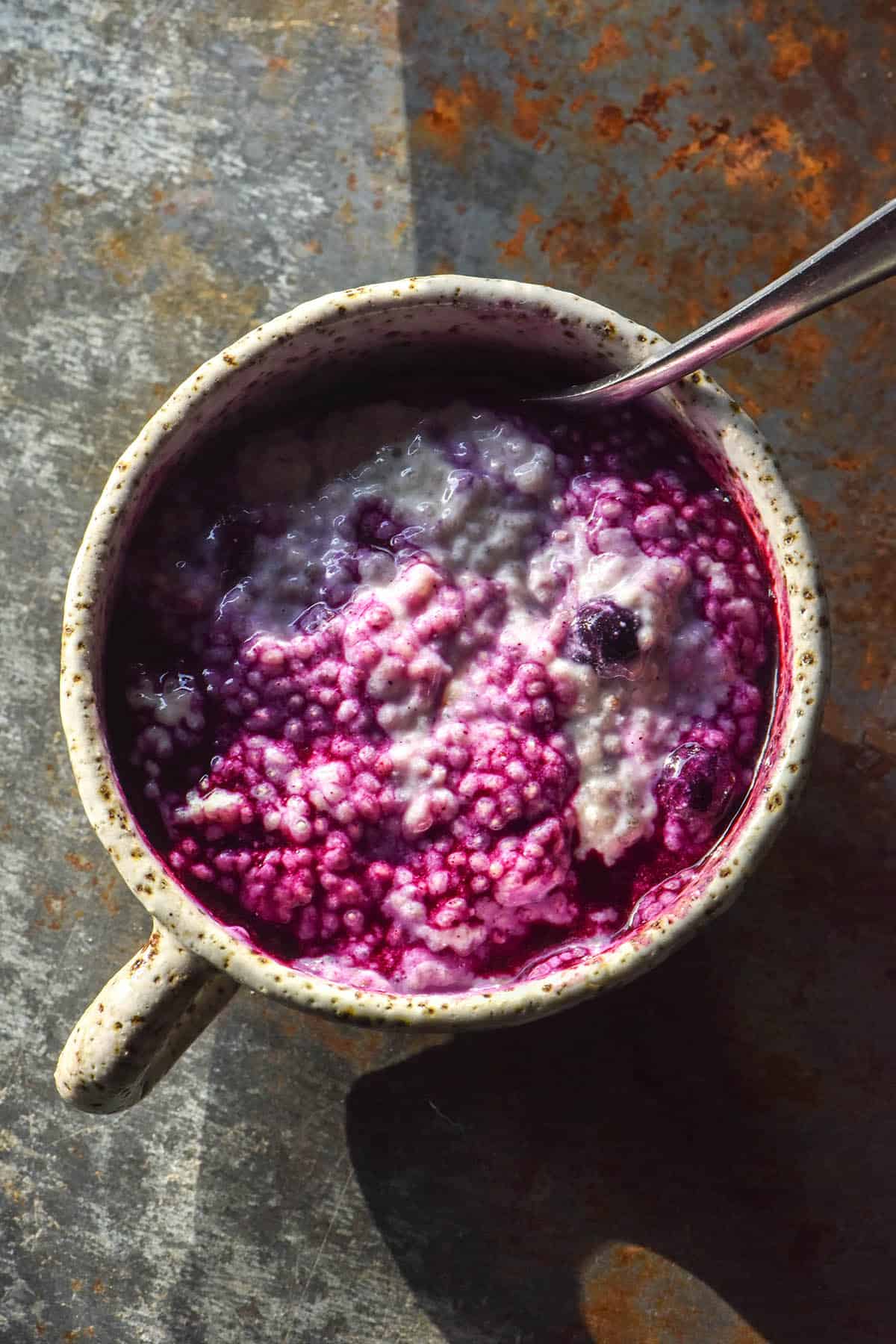An aerial image of a white speckled ceramic mug filled with a vibrant purple blueberry chia pudding. The mug sits atop a dark grey steel backdrop
