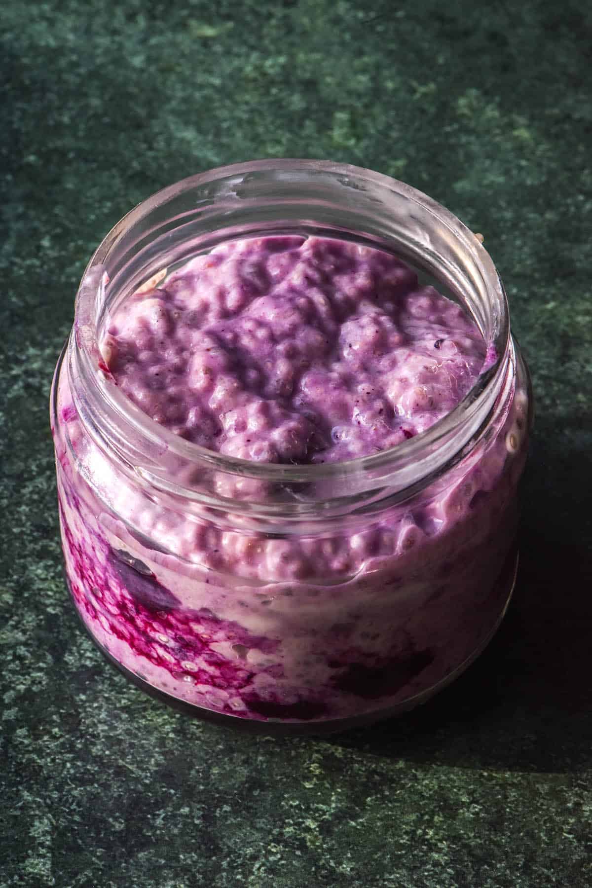 A side on image of a jar of low FODMAP chia pudding that has been swirled with defrosted frozen blueberries, giving it a vibrant purple colour. The chia pudding sits atop a dark green background