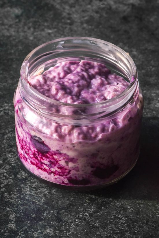 A side on image of a jar of low FODMAP chia pudding that has been swirled with defrosted frozen blueberries, giving it a vibrant purple colour. The chia pudding sits atop a dark grey background