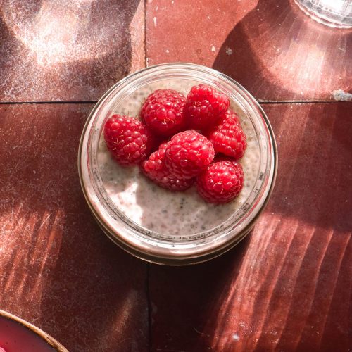 An aerial sunlit image of a chia pudding in a jar topped with raspberries. The jar sits atop a terracotta tile backdrop with two sunlit glasses of water at the top of the image.