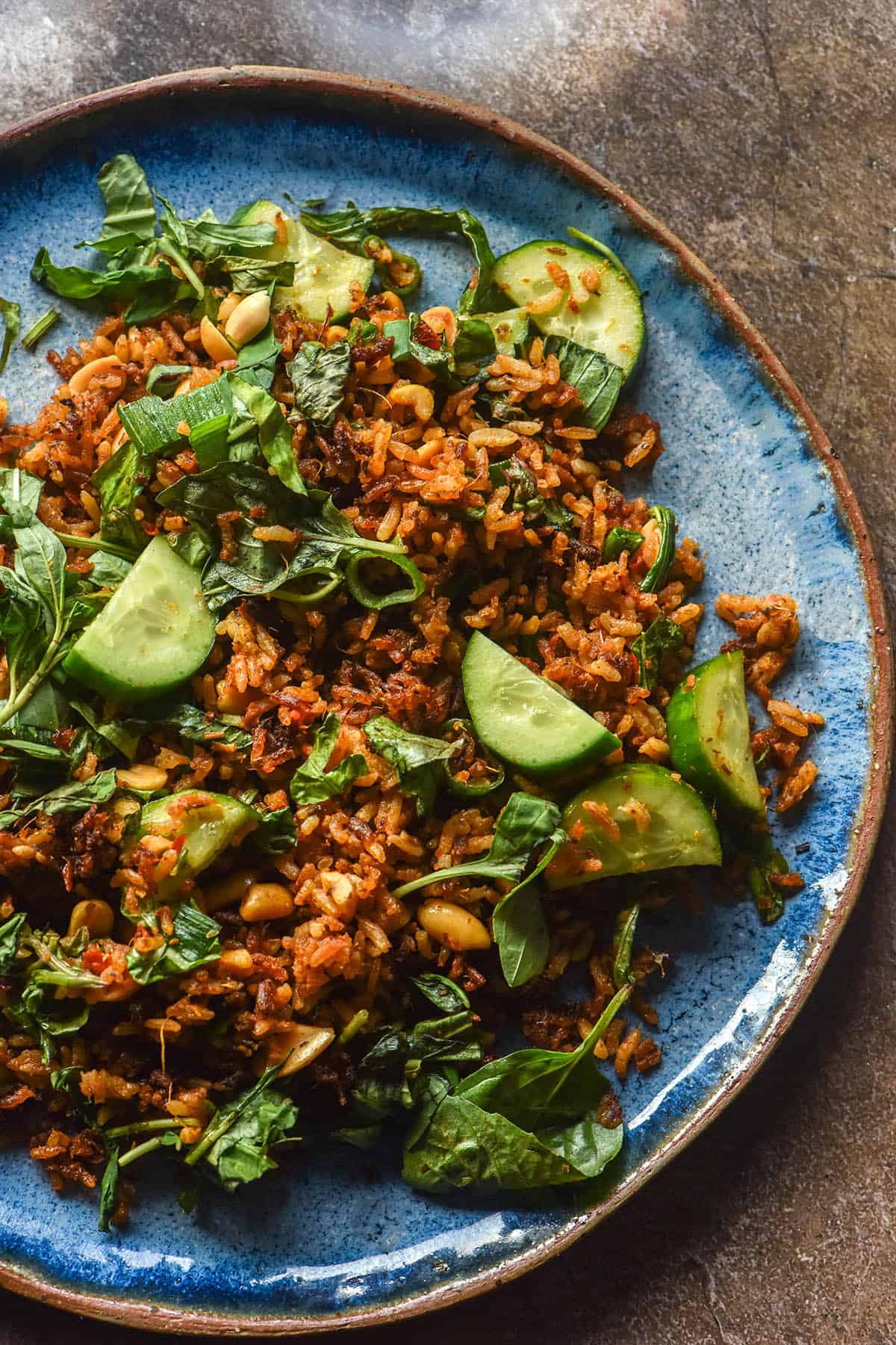 An aerial image of a bright blue ceramic plate topped with a crispy red rice salad, cucumbers, herbs and peanuts.