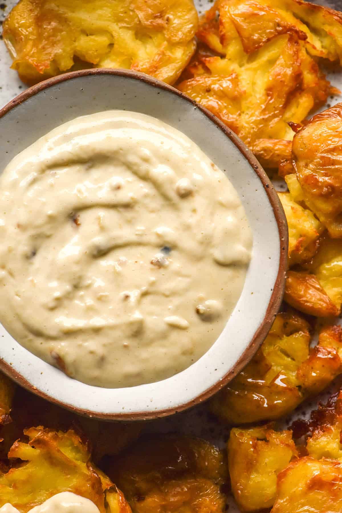 An aerial close up image of a white ceramic bowl filled with low FODMAP garlic aioli surrounded by crispy smashed potatoes