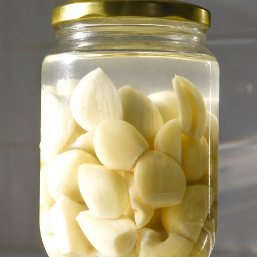A brightly lit image of a jar of pickled garlic on a white bench top against a white tiled backdrop