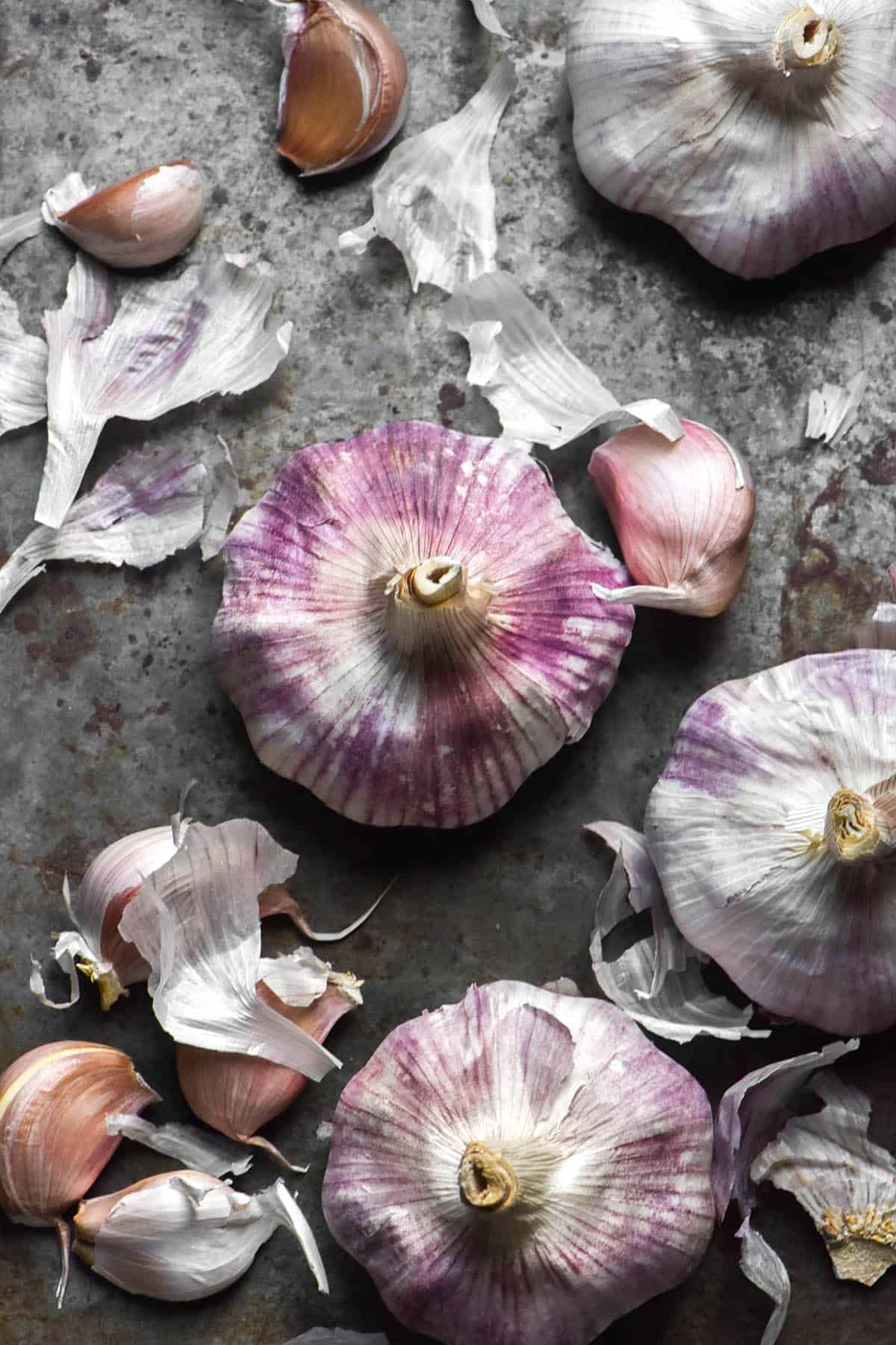 An aerial image of purple and white heads and cloves of garlic arranged casually on a dark steel backdrop 