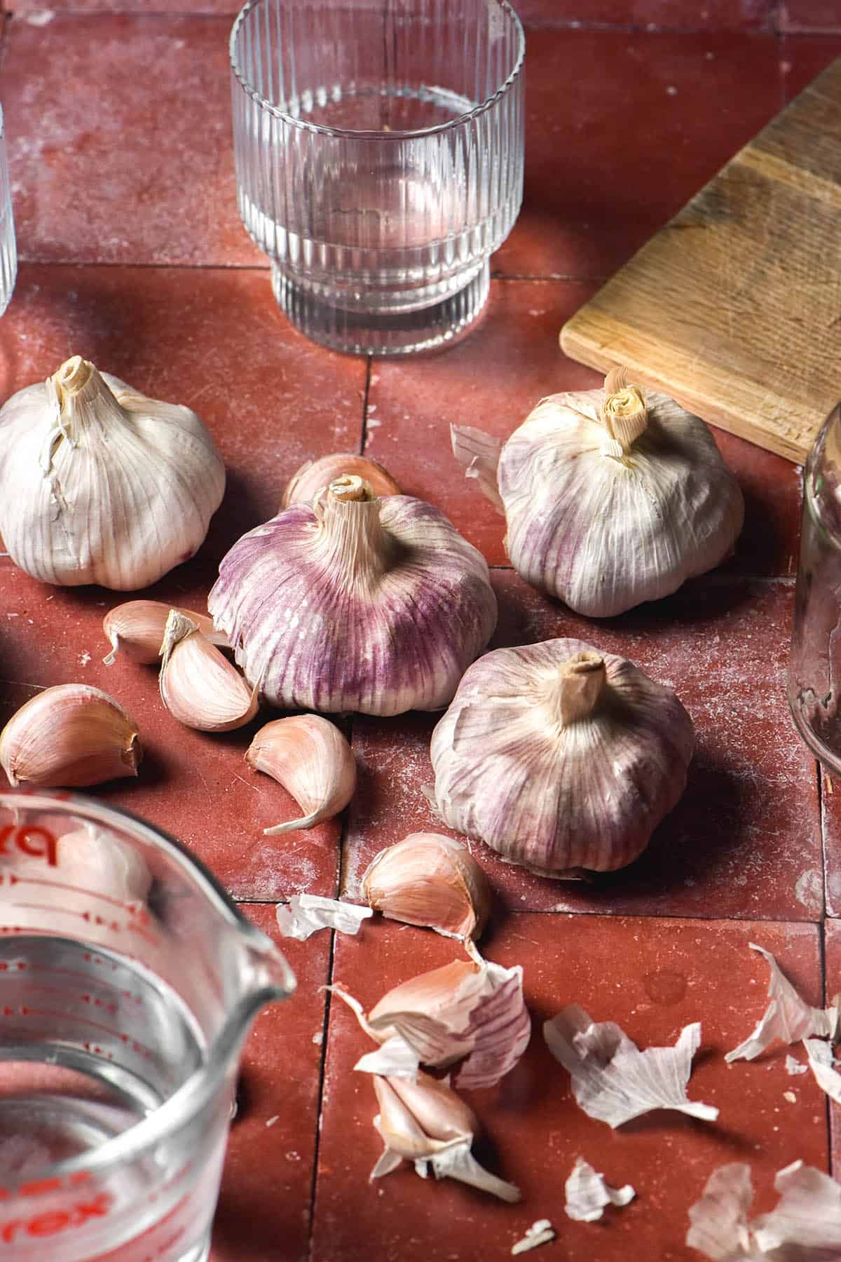 An aerial image of purple and white heads and cloves of garlic arranged casually on a terracotta tile backdrop. The garlic is surrounded by a chopping board, glasses of water and white vinegar for pickling