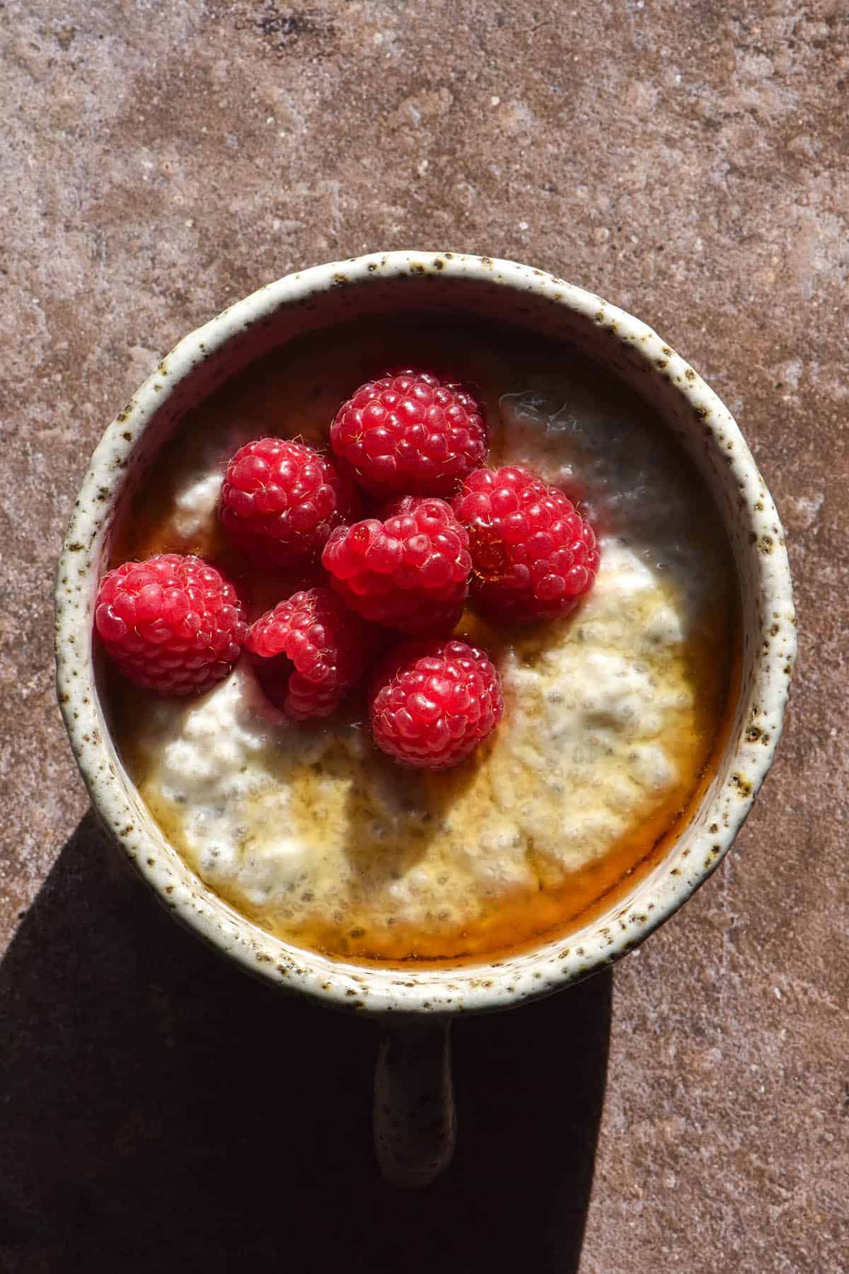 An aerial image of a white ceramic mug filled with low FODMAP vegan chia pudding and topped with fresh raspberries and maple syrup. The mug sits atop a dark grey concrete backdrop