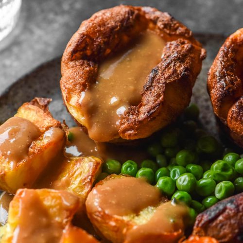 A side on image of a gluten free Yorkshire pudding filled with gravy and surrounded by vegetables. The food sits on a dark plate atop a dark backdrop and two glasses of water sit in the top left of the image