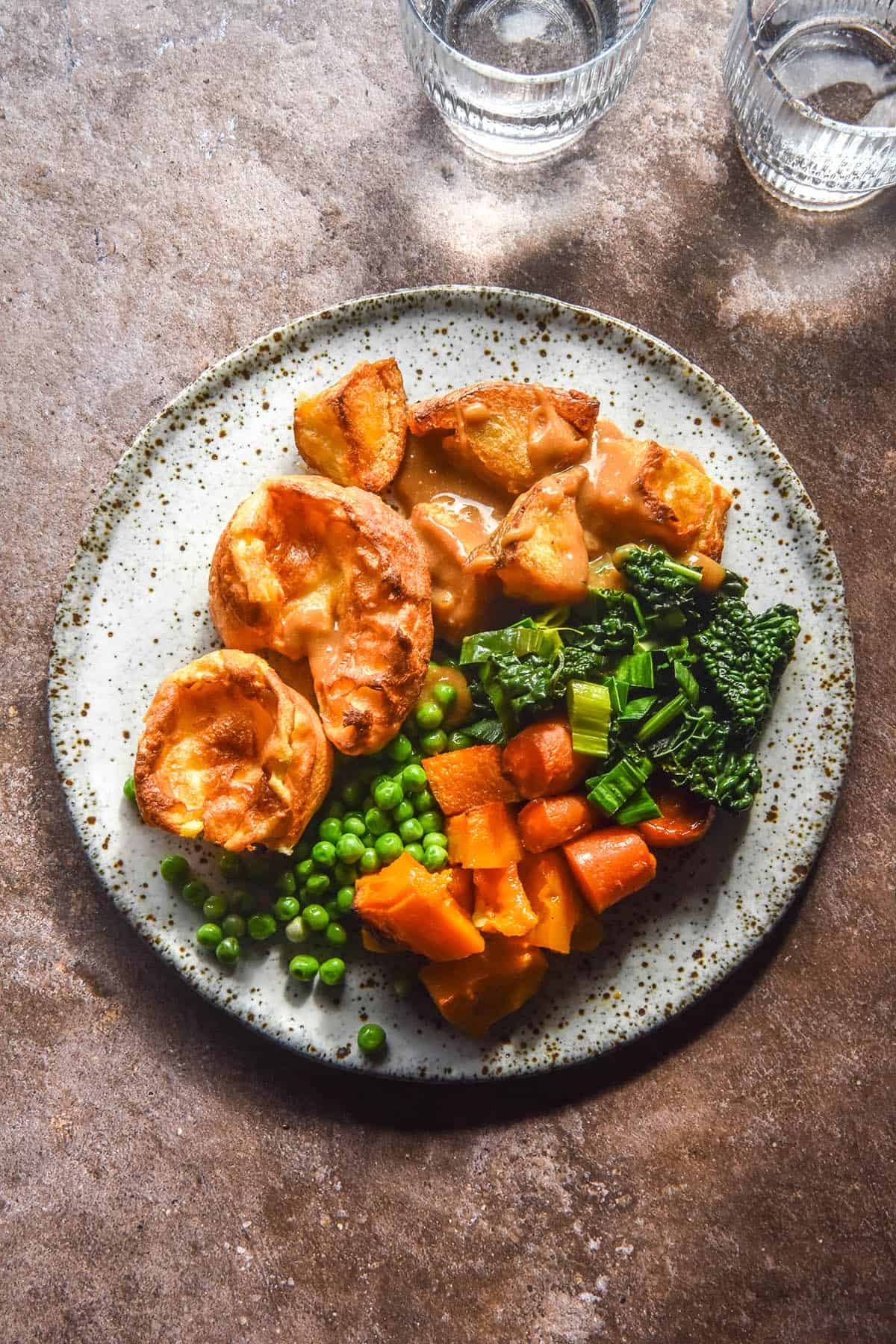 An aerial sunlit image of a white speckled ceramic plate of roast vegetables and gluten free Yorkshire puddings on a medium grey backdrop. Two glasses of water sit to the top right of the image