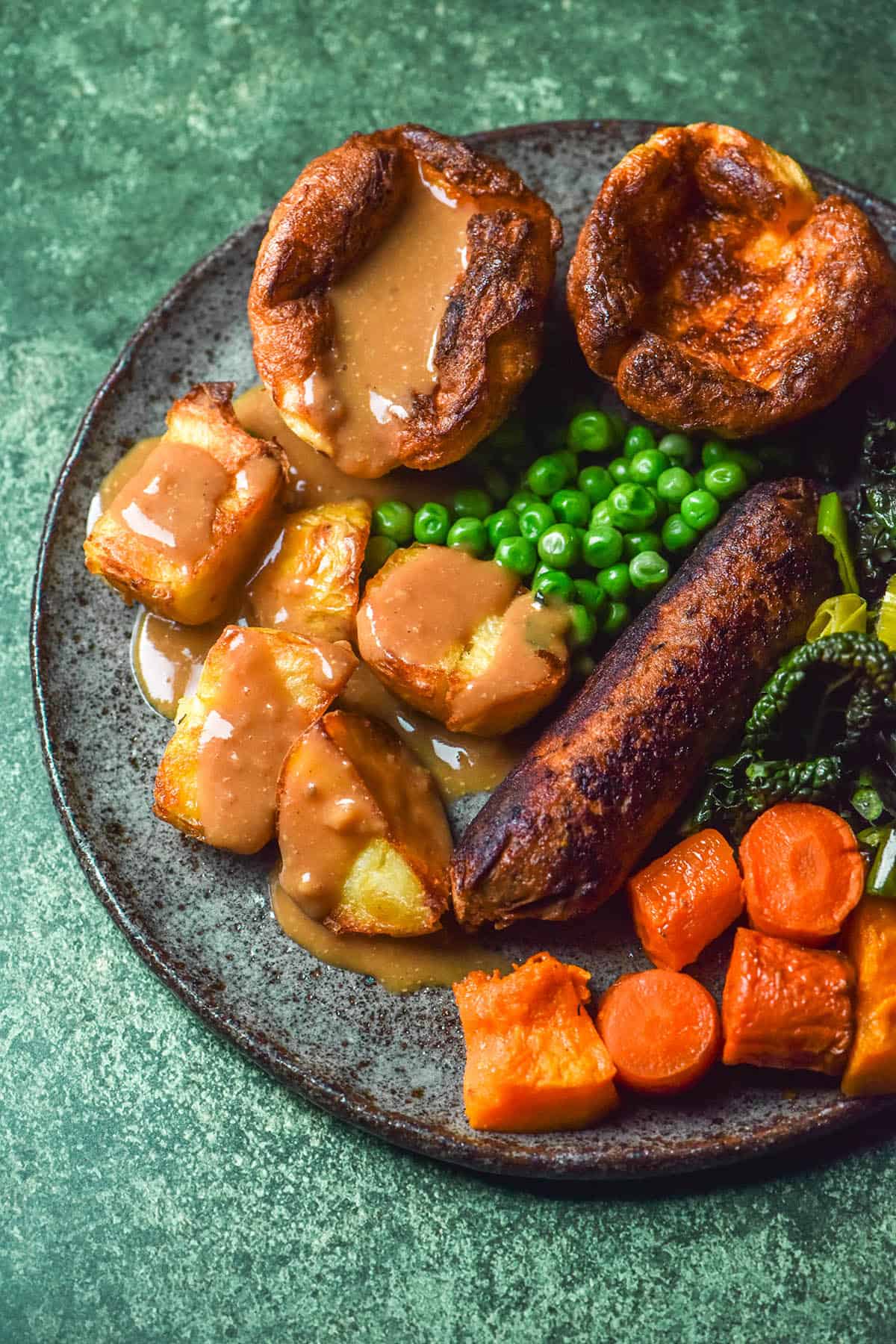 An aerial image of a dark blue ceramic plate topped with roast veggies, low FODMAP vegan sausage, gluten free Yorkshire puddings and gravy. The plate sits atop a bright green backdrop
