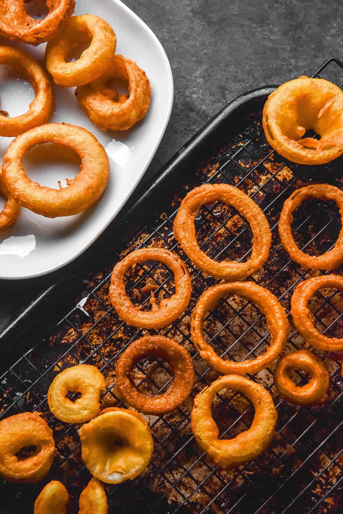 An aerial image of gluten free onion rings on a cooling rack and plate atop a dark backdrop