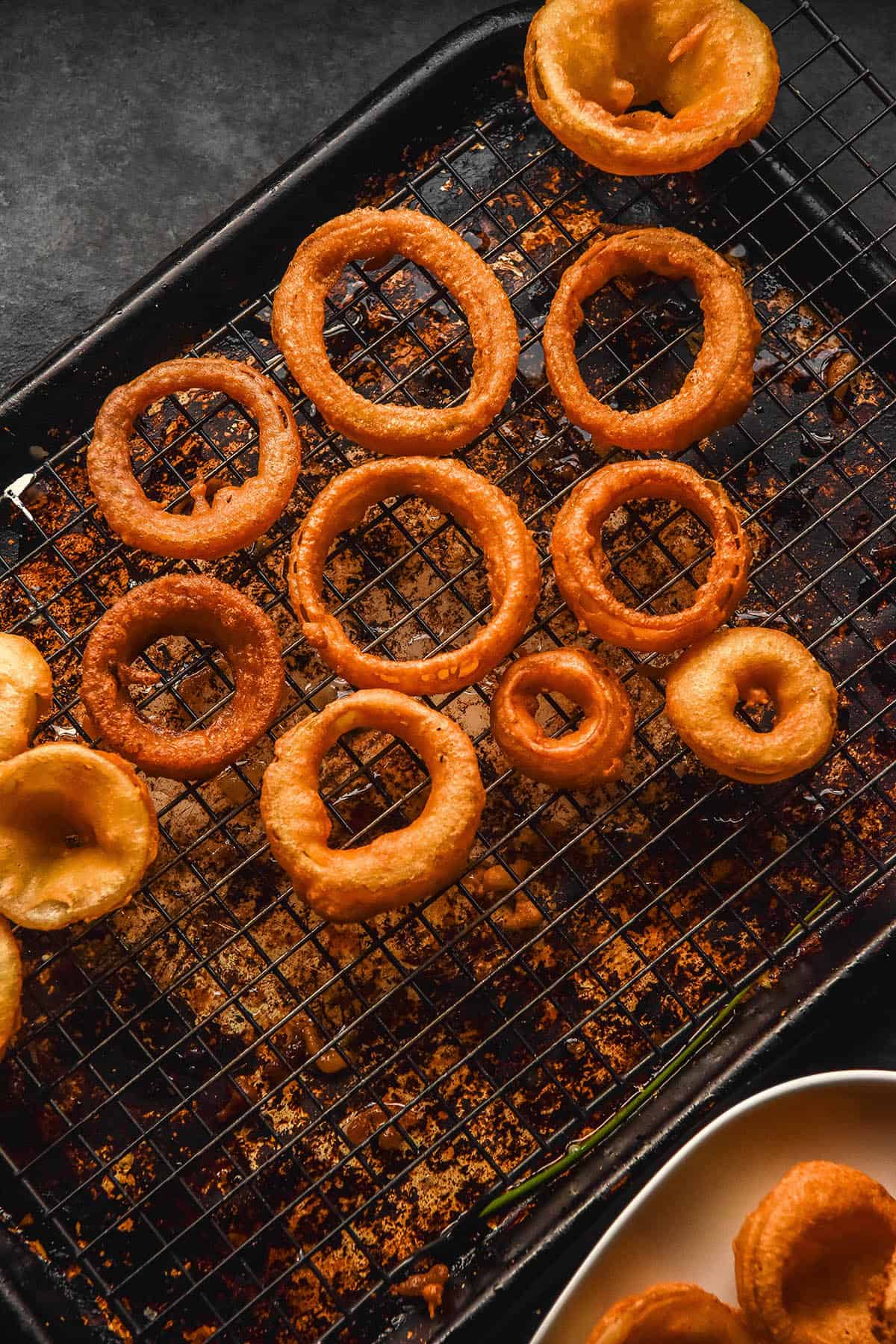 An aerial image of gluten free onion rings on a cooling rack and plate atop a dark backdrop