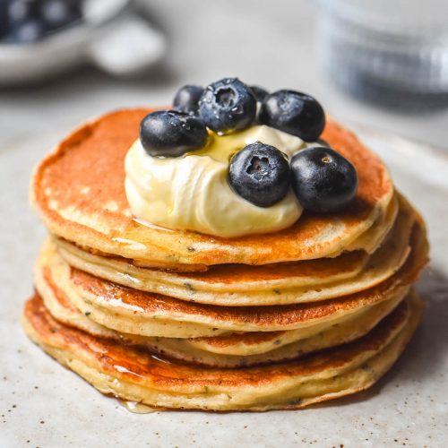 A side on image of gluten free protein pancakes without protein powder stacked and topped with yoghurt and blueberries. The pancakes sit on a small white plate atop a white marble table. A glass of water and a bowl of blueberries sit in the background