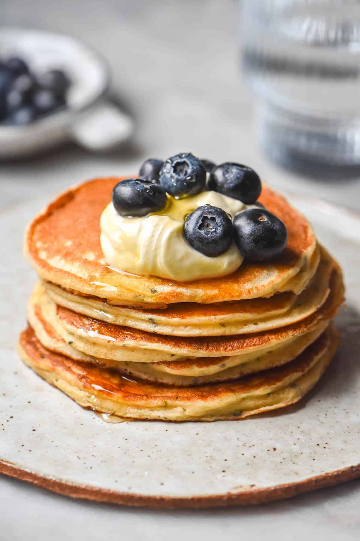 A side on image of gluten free protein pancakes without protein powder stacked and topped with yoghurt and blueberries. The pancakes sit on a small white plate atop a white marble table. A glass of water and a bowl of blueberries sit in the background