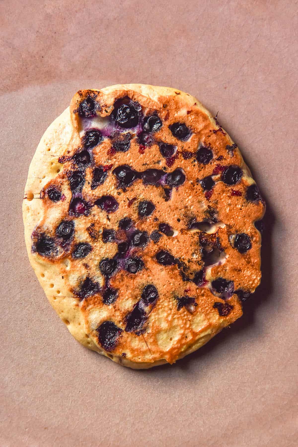 An aerial image of a gluten free protein pancake studded with lots of blueberries, sitting atop a pale pink ceramic plate