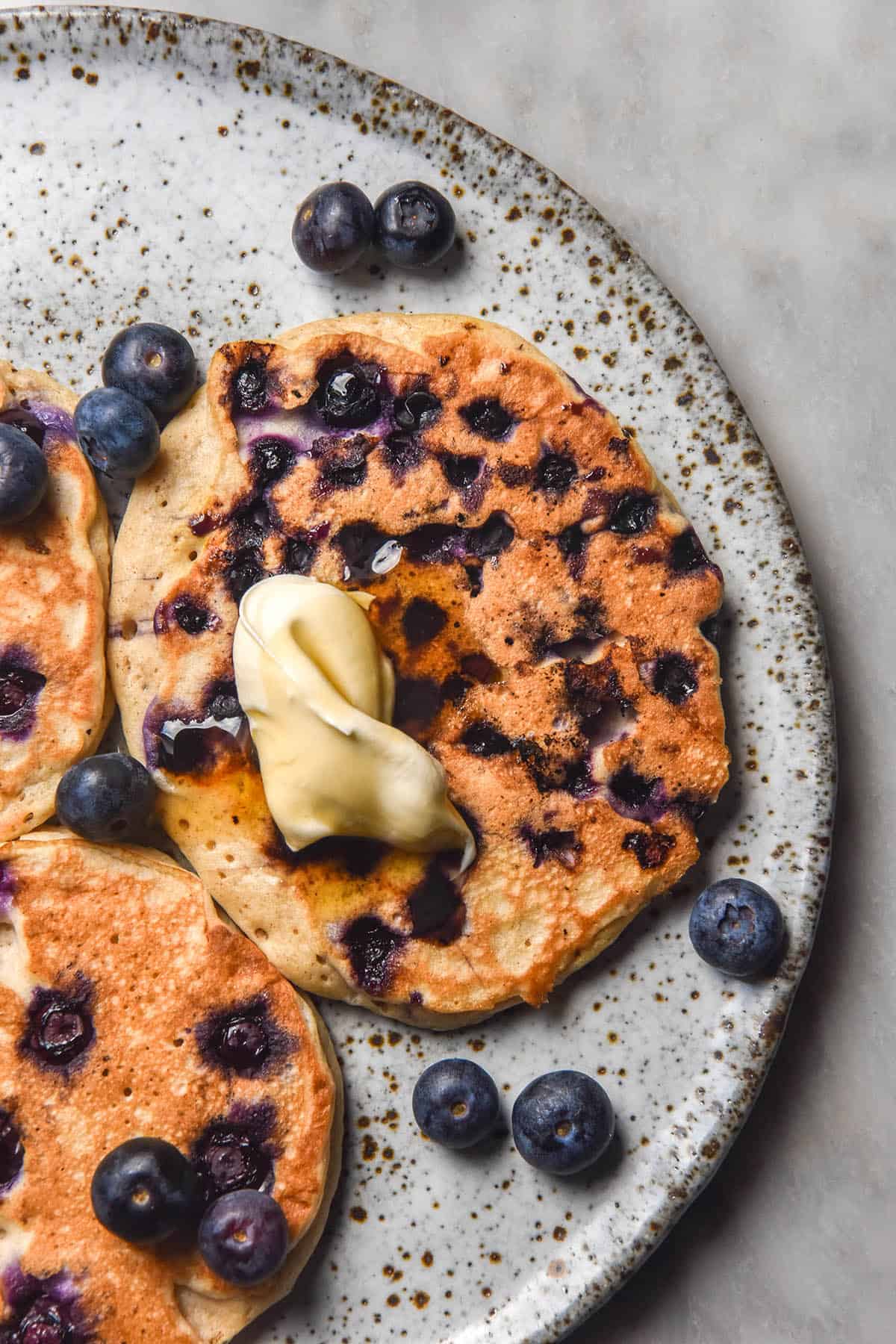 An aerial image of blueberry studded protein pancakes on a white speckled ceramic plate atop a white marble table