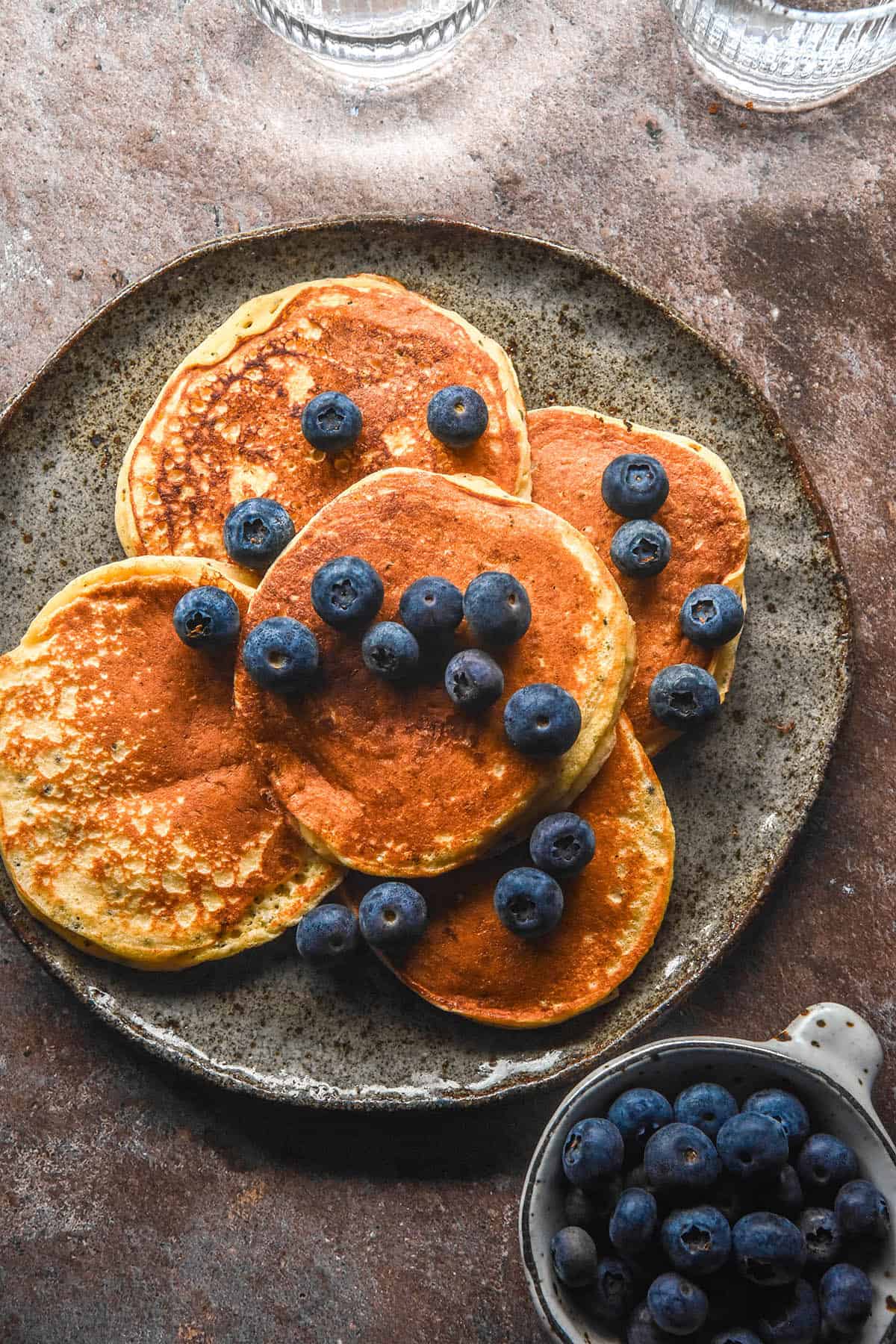 An aerial image of gluten free protein pancakes topped with blueberries on a dark blue plate atop a grey backdrop. A small white bowl of blueberries sits in the bottom right corner of the image and two glasses of water sit in the top of the image