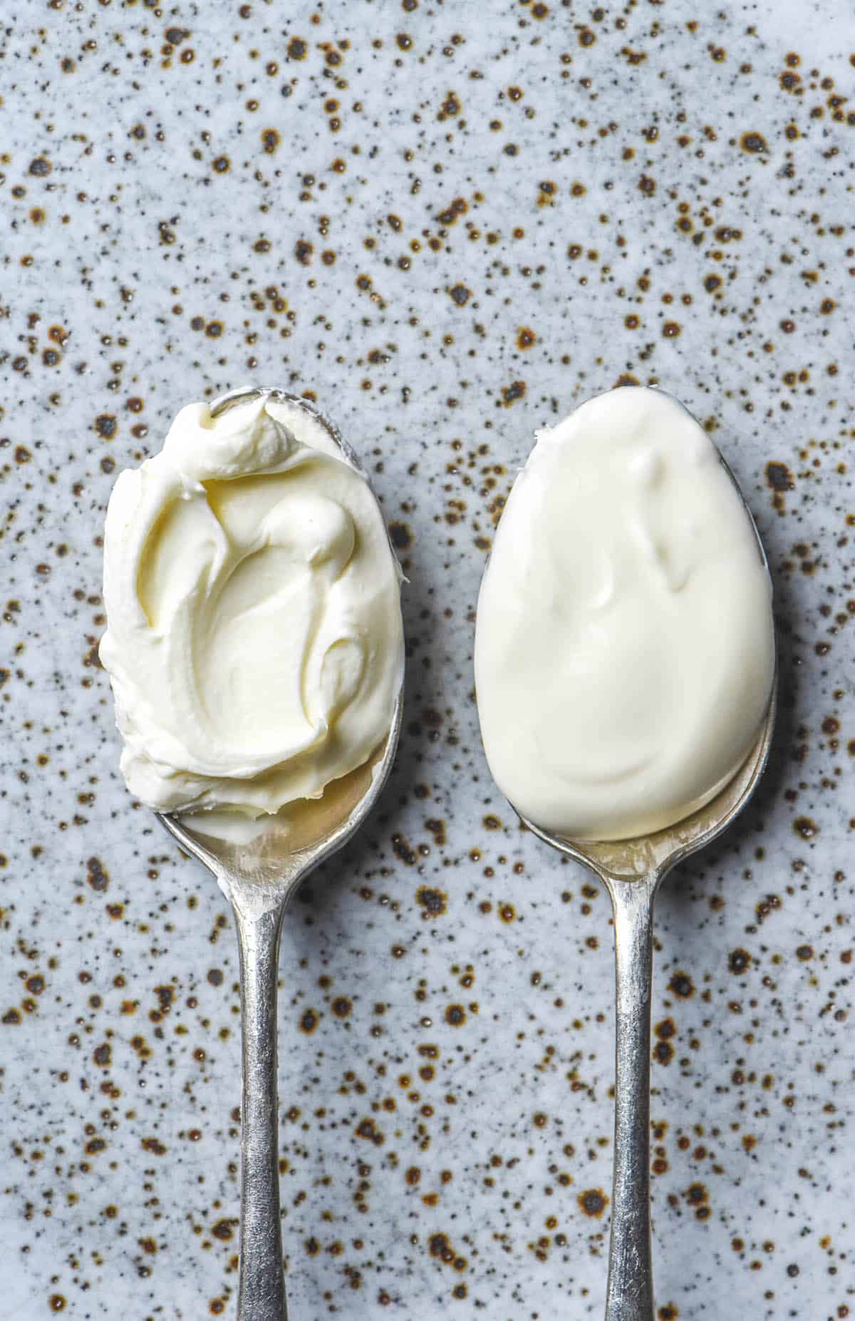 An aerial image of two spoons topped with lactose free sour cream atop a white speckled ceramic plate