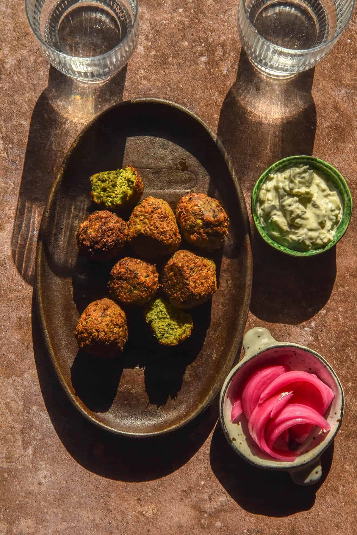 An aerial image of low FODMAP falafel on a dark grey platter atop a red grey backdrop. The falafel are surrounded by a small plate of low FODMAP tzatziki, two bowls of pickled red onions and a two sunlit glasses of water.