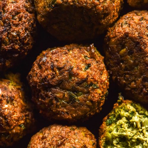 A macro overhead image of low FODMAP fried Falafel. The falafel are arranged in a tight knit pattern and the bottom left falafel is split open, revealing the bright green inside.