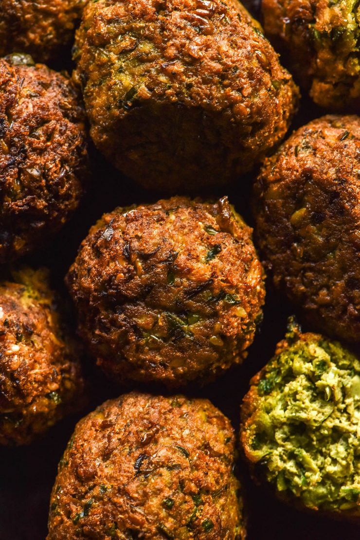 A macro overhead image of low FODMAP fried Falafel. The falafel are arranged in a tight knit pattern and the bottom left falafel is split open, revealing the bright green inside.