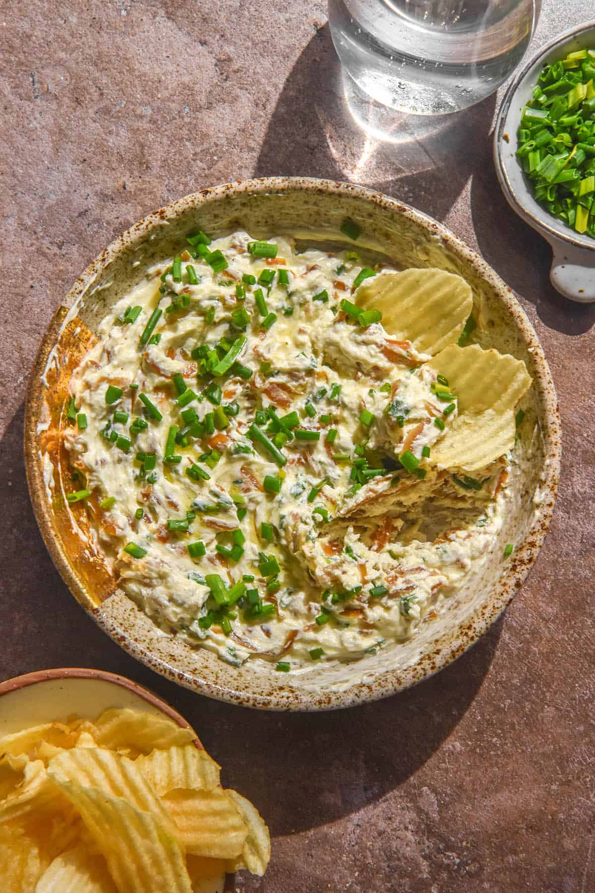 An aerial image of a speckled ceramic bowl filled with low FODMAP French Onion Dip. The dip has been topped with lots of chopped chives and some crinkled cut chips sit in the top right corner of the bowl. The bowl sits atop a medium grey backdrop and extra bowls and glasses of water surround the bowl of dip.