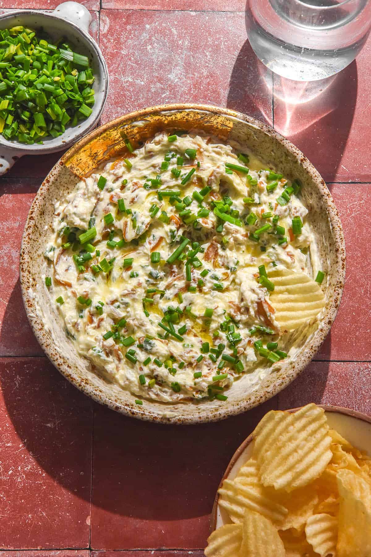 An aerial image of a bowl of low FODMAP French onion dip atop a terracotta tile backdrop. The dip is surrounded by a sunlit water glass, a bowl of chives and a bowl of crinkle cut chips