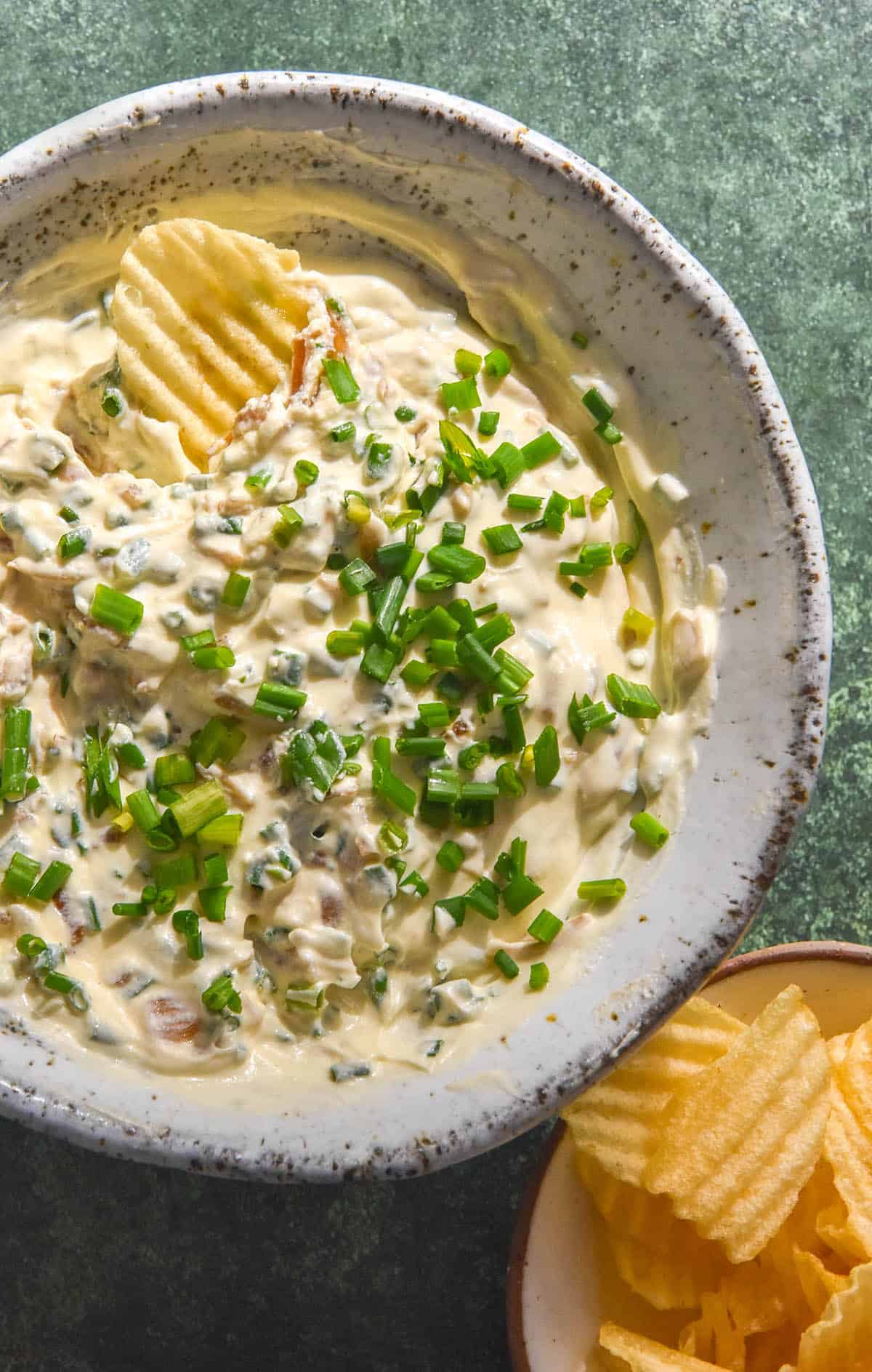 An aerial image of a white speckled ceramic bowl filled with low FODMAP French Onion Dip. The dip is topped with chopped chives and a crinkle cut chip dips into the top left corner