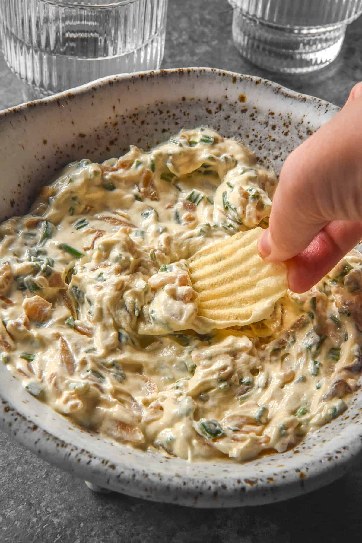 A side on image of a white speckled ceramic bowl filled with low FODMAP French Onion Dip. A hand dips into the bowl from the right side with a crinkled cut chip. Two glasses of water sit in the background of the image.