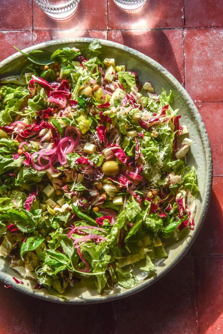 A sunlit aerial image of a low FODMAP chopped salad on a ceramic serving platter atop a terracotta backdrop. Two sunlit glasses of water sit in the top of the image.