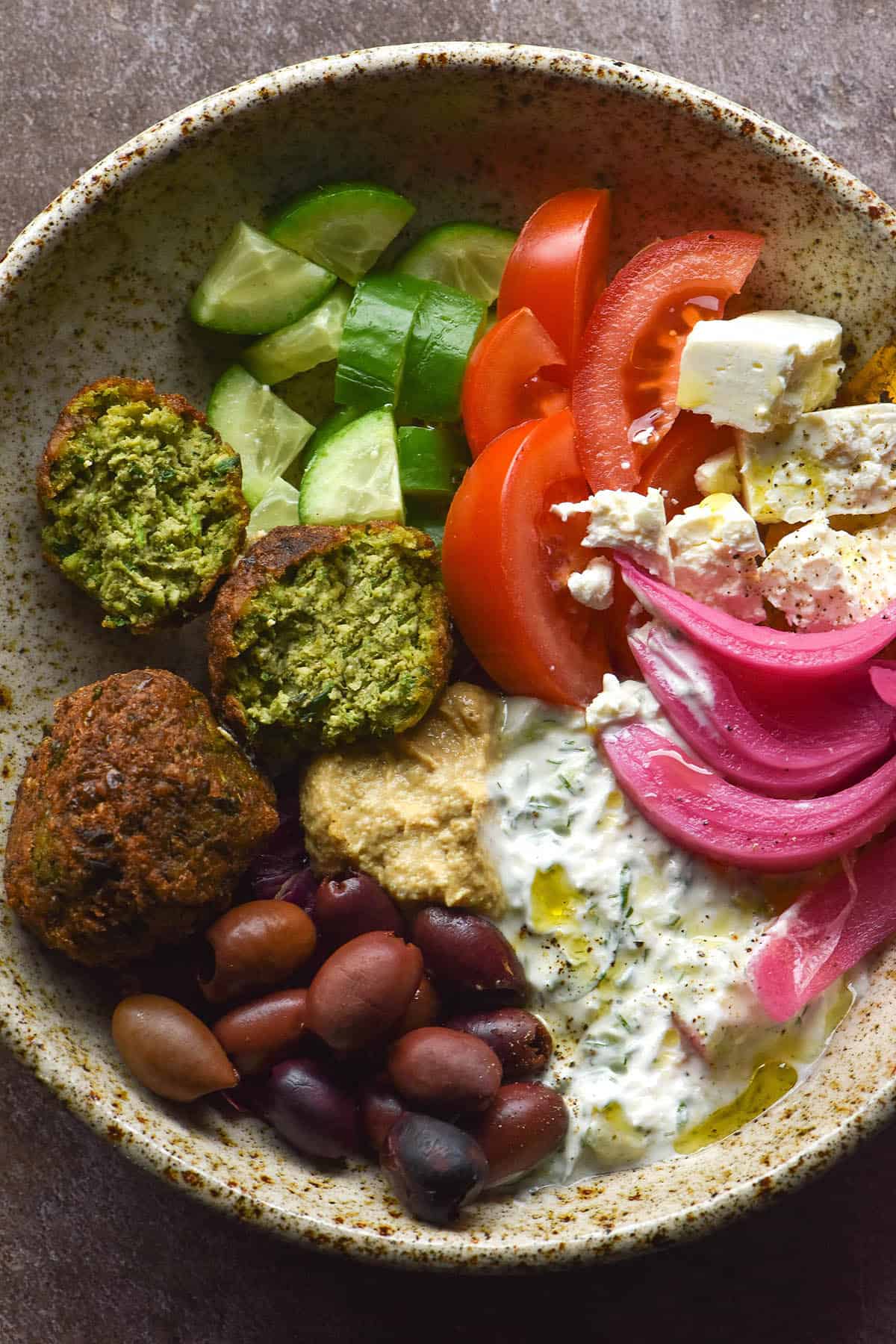 An aerial image of a low FODMAP falafel bowl filled with edamame falafel, tomatoes, cucumber, pickled red onion, feta, low FODMAP hummus and low FODMAP tzatziki.