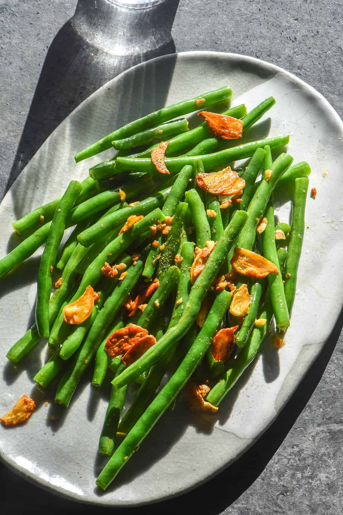 A brightly lit aerial image of low FODMAP green beans topped with low FODMAP garlic chips. The beans sit on a white oval plate atop a dark blue backdrop
