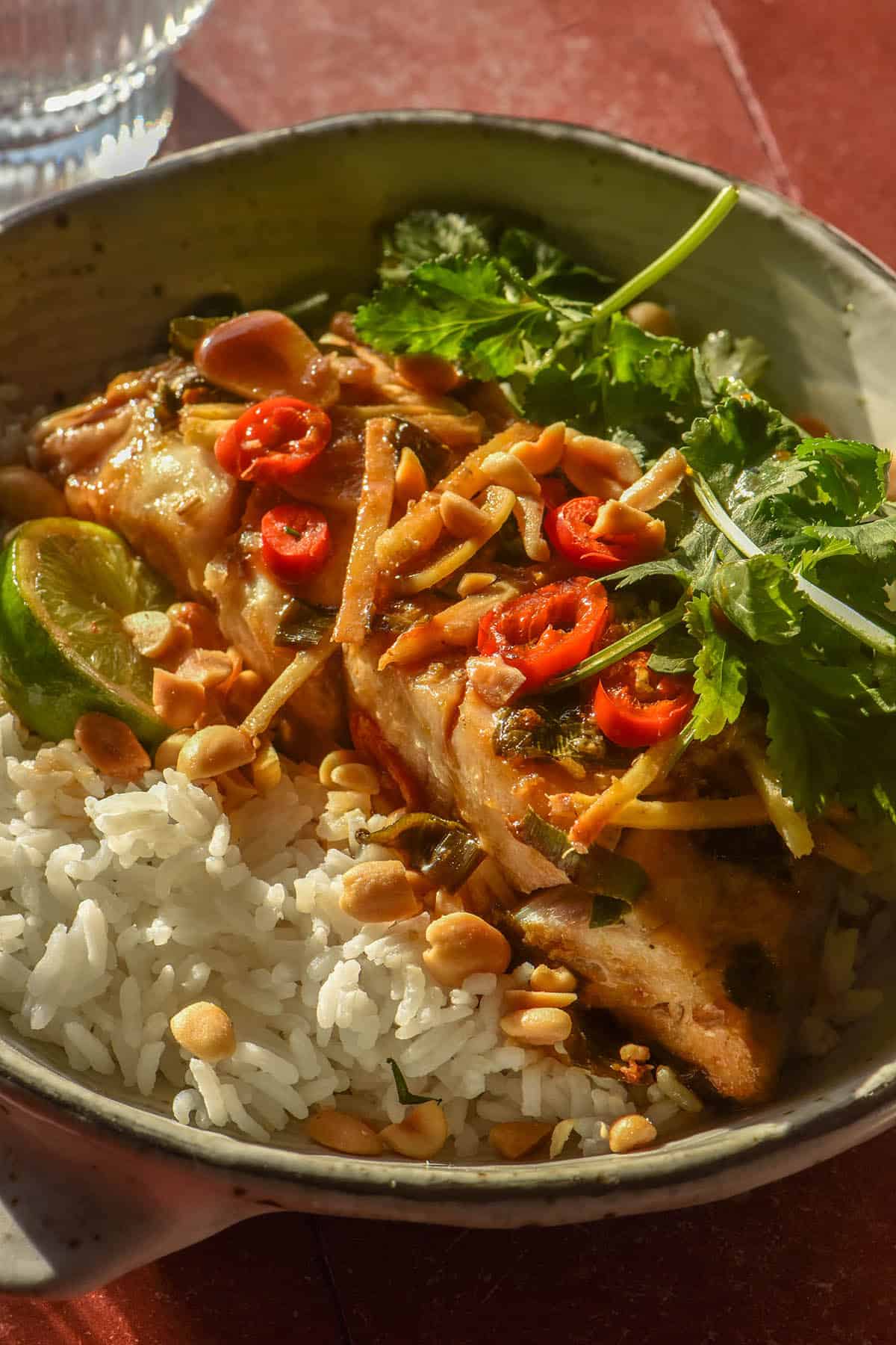 A close up image of a bowl filled with rice and low FODMAP Asian baked fish topped with chilli, coriander and peanuts. The bowl sits atop a terracotta tile backdrop and a glass of water sits to the top left of the image