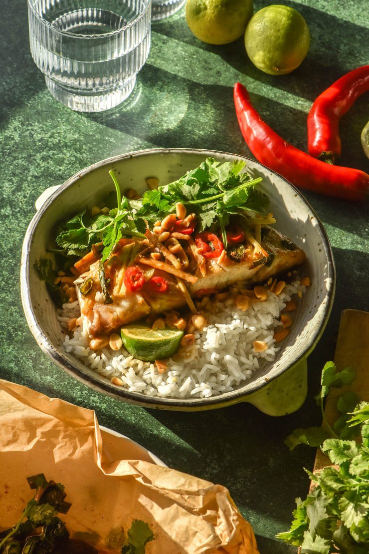 A side on image of Asian baked barramundi on a bed of rice in a white ceramic bowl. The fish is topped with coriander and red chilli and the bowl sits atop an olive green backdrop. It is surrounded by ingredients for the fish and a sunlit glass of water.