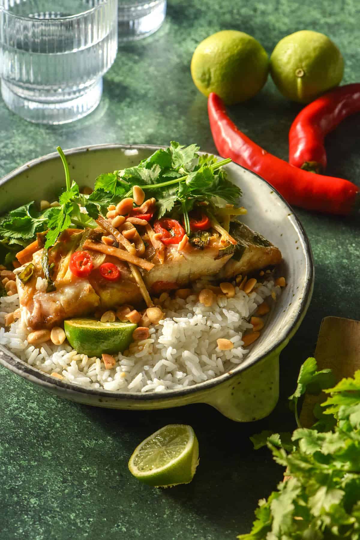 A side on image of Asian baked barramundi on a bed of rice in a white ceramic bowl. The fish is topped with coriander and red chilli and the bowl sits atop an olive green backdrop. It is surrounded by ingredients for the fish and a sunlit glass of water.