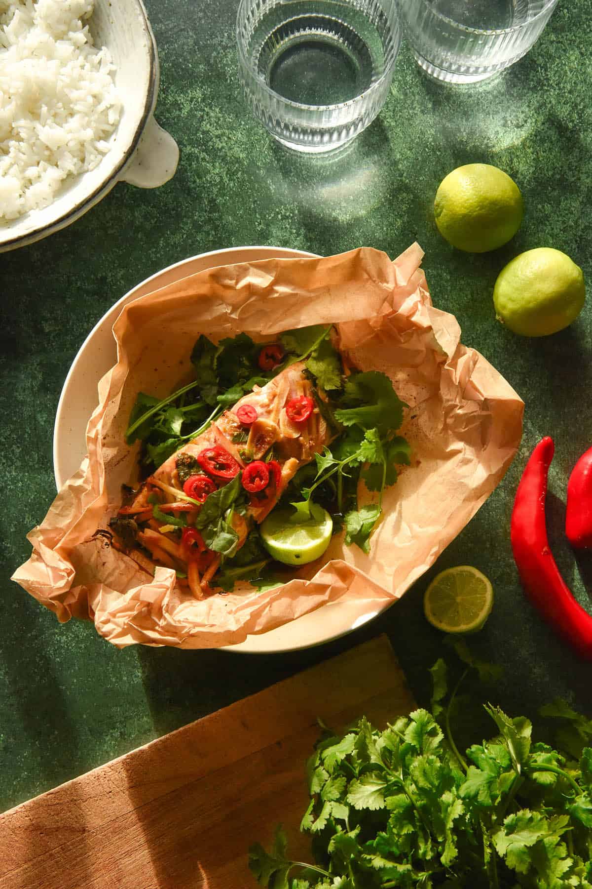 An aerial image of a piece of fish baked in a baking paper parcel with Asian inspired flavours. The fish sits in the centre of the image and is surrounded by ingredients used to flavour the dish, glasses of water and white rice for serving