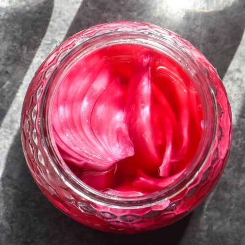 An aerial image of a jar of low FODMAP pickled onions atop a grey backdrop with two sunlit glasses of water in the top of the image. The onions and brine are a vibrant pink purple colour which pops against the grey backdrop.