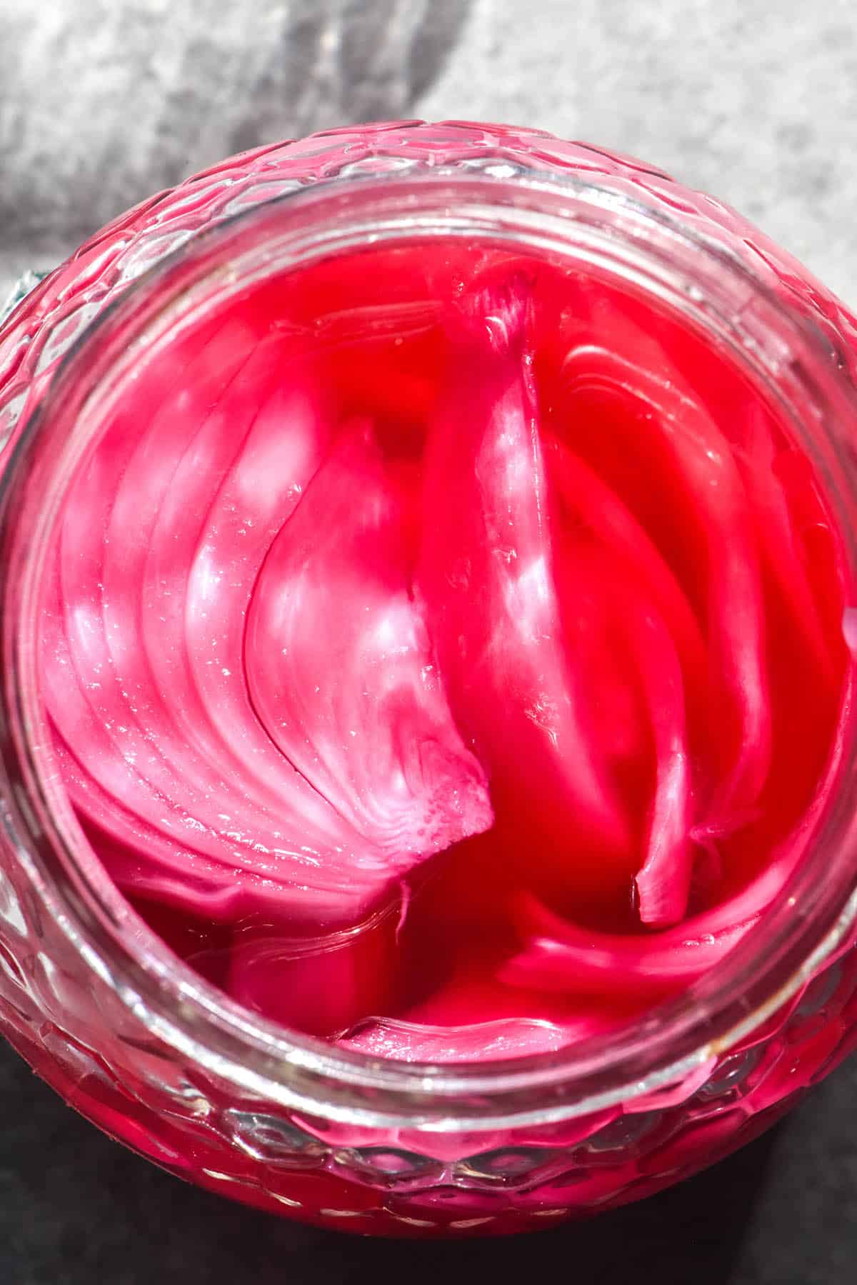 A sunlit macro aerial close up of a jar of pickled red onions against a light grey backdrop