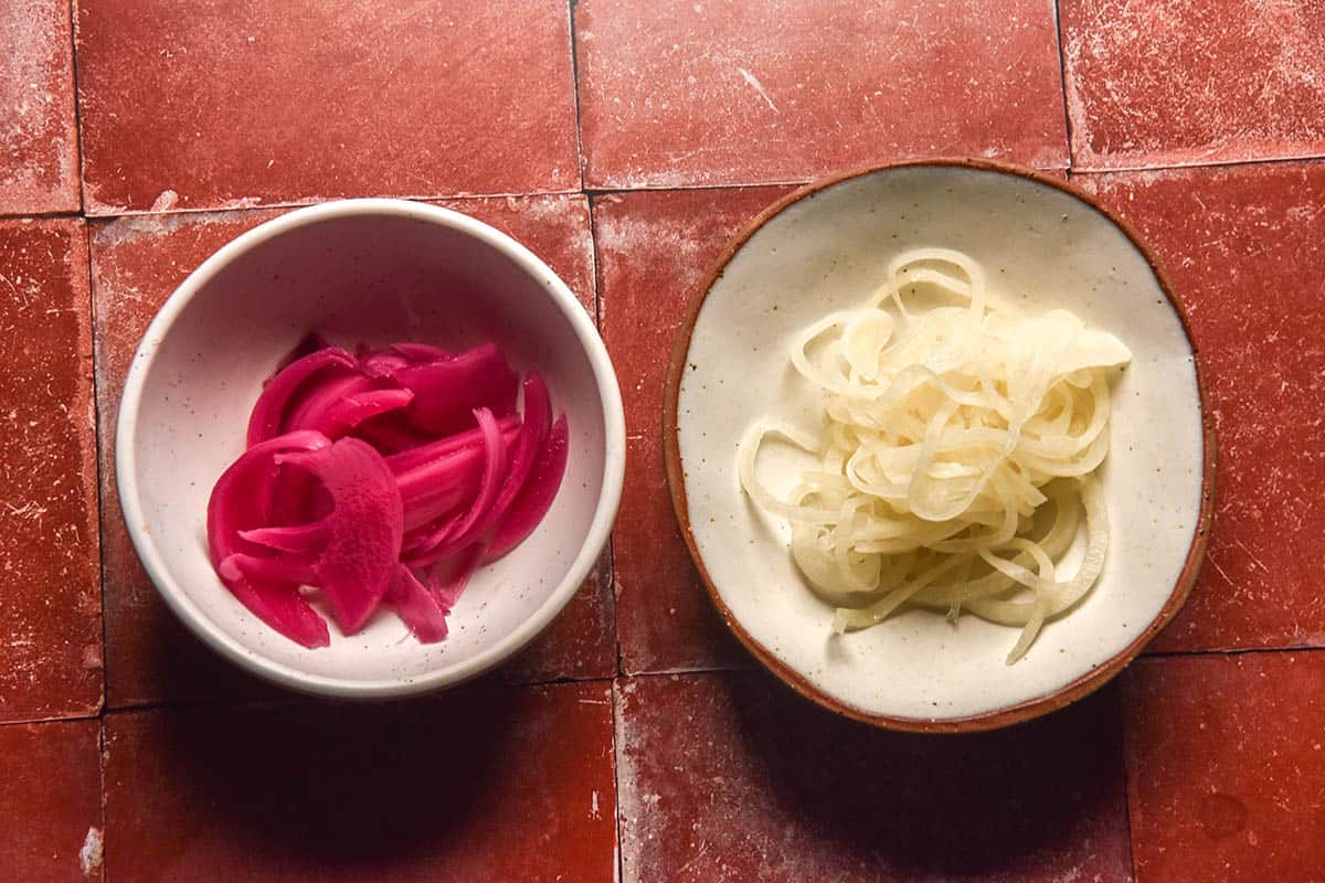 An aerial image of two bowls of pickled onion on a terracotta tile backdrop. 