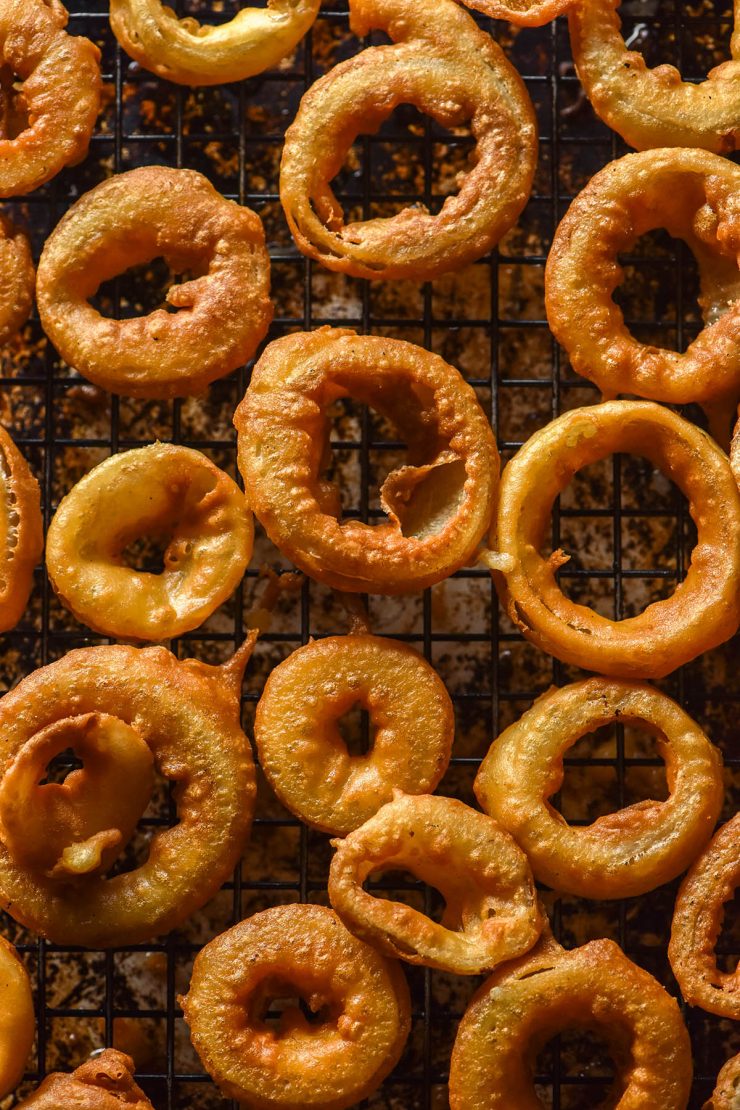 An aerial image of low FODMAP onion rings on a baking rack atop a dark brown baking tray. The onion rings are crispy and golden brown.
